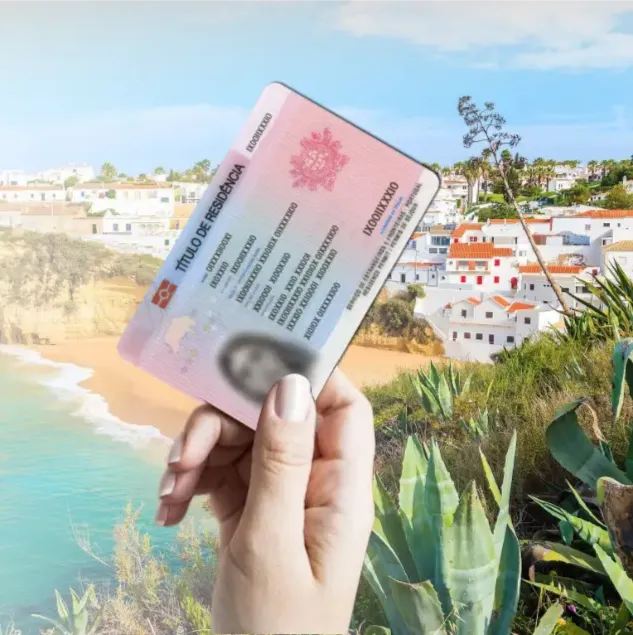 A Portuguese residence card (Título de Residência) held up against a picturesque background of coastal houses and lush vegetation, typical of Portugal’s scenic seaside areas.