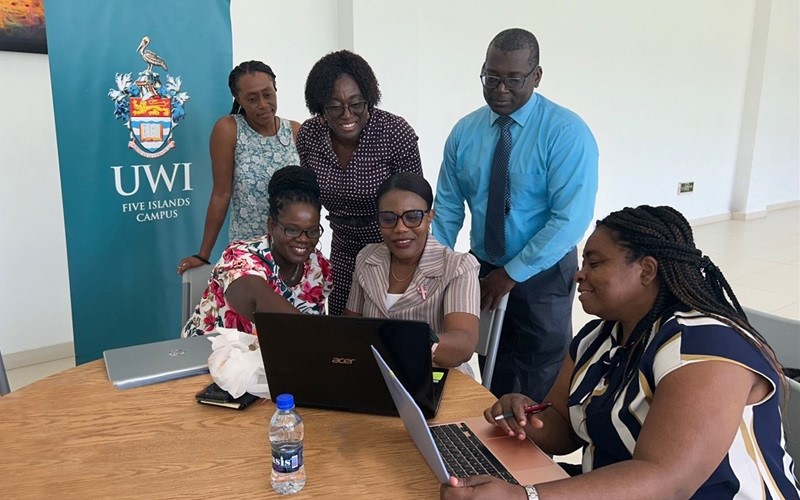 Group of professionals collaborating around laptops, with a UWI Five Islands Campus banner in the background.