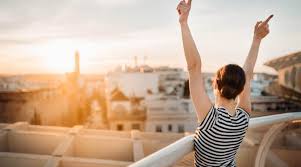 Woman standing with arms raised, overlooking a cityscape during sunset.