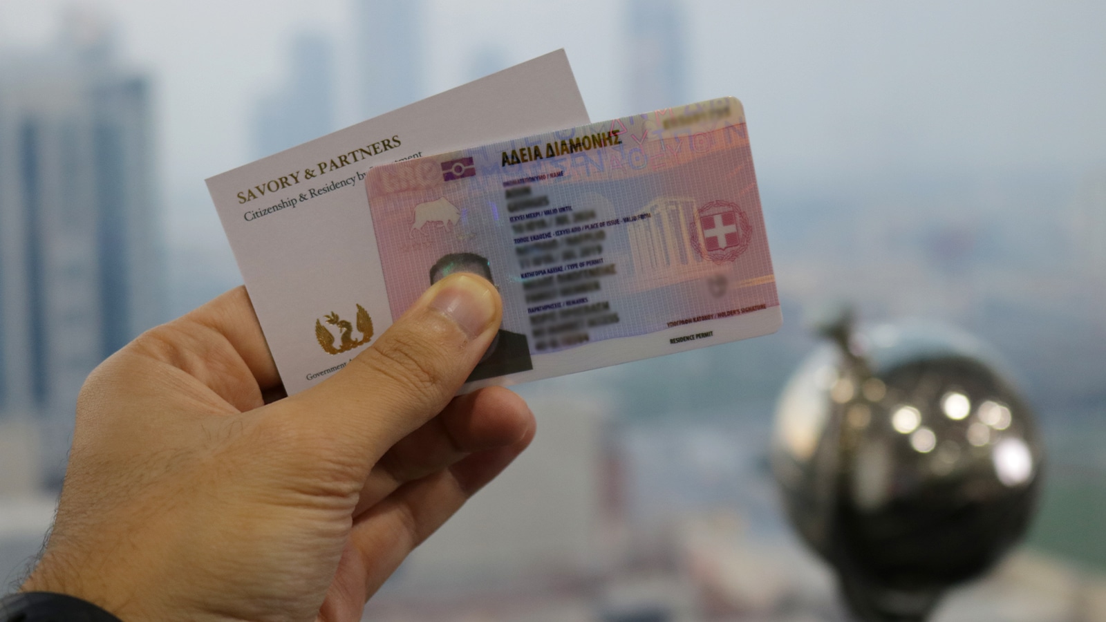 Hand holding a Greek residence permit card alongside a business card from Savory & Partners, with a blurred cityscape in the background.