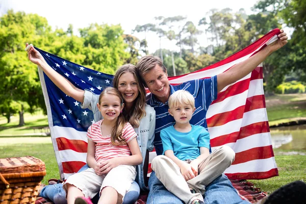 A happy family sitting outdoors on a picnic blanket, holding the American flag, with a park and pond in the background.