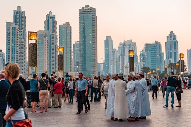 A bustling city scene with a diverse crowd of people gathered in a plaza, set against a backdrop of modern skyscrapers.