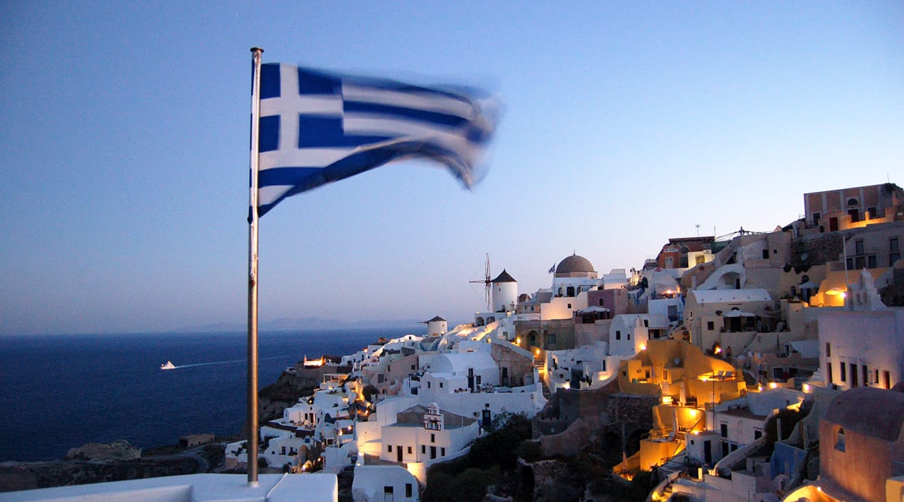 Greek flag waving above a scenic view of Santorini with white-washed buildings, windmills, and the Aegean Sea at dusk.